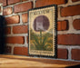 A metal sign with a purple flower and the word "ALLIUM" printed on it, mounted on a brick wall.