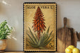 A framed aloe vera plant with a vintage postage stamp design, placed on a shelf next to a wooden cutting board and some lemons.