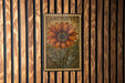 A framed image of a large orange flower with green leaves, mounted on a wooden wall.