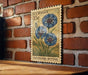 A vintage U.S. postage stamp featuring a blue flower design, mounted on a brick wall.
