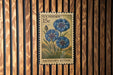 A U.S. postage stamp featuring a blue flower, mounted on a wooden wall.