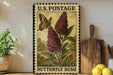 A vintage U.S. postage stamp featuring a butterfly and a flowering bush, with the text "Butterfly Bush" printed on it. The stamp is displayed on a shelf next to a wooden cutting board and a potted plant.