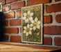A framed botanical illustration of clematis flowers is displayed on a brick wall, with a wooden table below it.