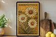 A framed illustration of three daisies is displayed on a shelf, accompanied by a wooden cutting board and a potted plant.