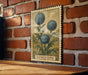 A vintage-style metal stamp depicting a thistle flower is mounted on a brick wall.