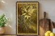 a framed print of a large orchid flower, with a vintage-style stamp design, placed on a shelf next to a wooden cutting board and some potted plants.