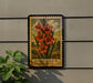 A framed illustration of a Gladiolus flower is displayed on a wall, with a plant to the left of the frame.