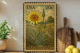 A framed postage stamp depicting a yellow flower with green leaves, placed on a shelf next to a wooden cutting board and a potted plant.