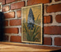 A framed illustration of a blue flower is hanging on a brick wall, with a wooden table below it.