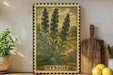 a framed illustration of blue flowers, with the word "HYSOP" written below the illustration. The frame is placed on a wooden surface, with a potted plant to the left and a wooden cutting board to the right.