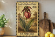 A vintage-style postage stamp featuring a pink flower, with the text "Lady's Slipper (Pink)" and "20c" printed on it. The stamp is displayed on a shelf next to a wooden cutting board and a potted plant.