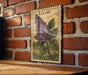 A framed illustration of a purple lilac flower is displayed on a brick wall.