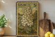 A framed illustration of a white lilac flower on a wooden shelf, with a vintage postage stamp design.