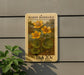 A framed postage stamp featuring a painting of yellow flowers on a green plant, mounted on a wall.