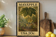 A framed stamp depicting a plant with a large leaf and a flower, placed on a shelf next to a wooden cutting board and some lemons.