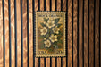 A vintage-style postage stamp from the United States, featuring a floral design with white flowers and green leaves, mounted on a wooden wall.