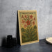a vintage-style stamp or postcard with a floral illustration, placed on a dark surface next to some black camera lenses.