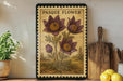 a framed illustration of a pasque flower on a shelf, with a potted plant to the left and a wooden cutting board to the right.