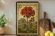 A framed illustration of a red gerbera daisy on a wooden shelf, with a kitchen cutting board and lemons nearby.