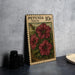 A wooden postcard with a floral illustration and text, placed on a table next to some black camera lenses.