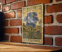 A vintage-style postage stamp featuring a blue wild blue flower is displayed on a brick wall.