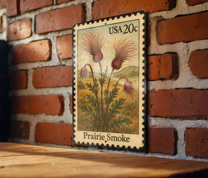 A framed postage stamp depicting a field of flowers, specifically a prairie smoke flower, is displayed on a brick wall.