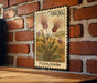 A framed postage stamp depicting a field of flowers, specifically a prairie smoke flower, is displayed on a brick wall.