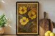 A framed stamp depicting yellow flowers is displayed on a shelf, surrounded by kitchen utensils and a potted plant.