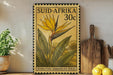 A vintage postage stamp featuring a yellow flower with green leaves, placed on a wooden surface next to a wooden cutting board and a potted plant.