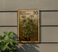 a wooden postcard-style sign with a floral design, mounted on a wall next to a plant.