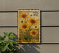 A postage stamp featuring a vibrant illustration of yellow flowers, with the word "TICKSEED" printed below. The stamp is affixed to a gray wall, and there is a small green plant to the left of the stamp.