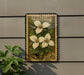 A vintage U.S. postage stamp featuring a trillium flower is mounted on a wall, with a plant to the left.