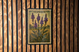 a framed illustration of purple flowers on a wooden wall.