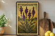 a framed illustration of purple flowers on a wooden shelf, with a wooden cutting board and a potted plant nearby.