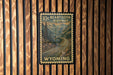 A vintage-style postage stamp depicting a scenic highway in Wyoming, mounted on a wooden wall.