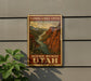 A framed vintage postage stamp depicting a scenic mountain landscape with a river, hanging on a wall next to a plant.