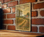 A vintage-style postage stamp depicting a rural road leading through a countryside landscape, mounted on a brick wall.