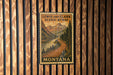 A vintage-style metal sign with a scenic mountain landscape and a winding road, mounted on a wooden wall.