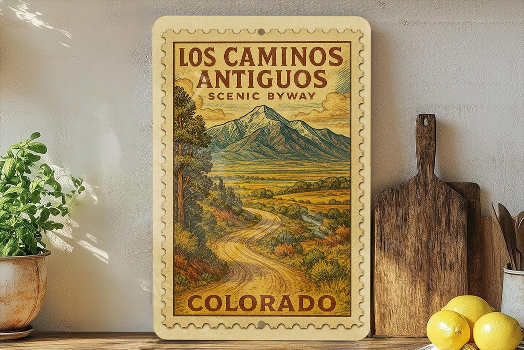 A framed vintage-style poster of the scenic route in Los Caminos Antiguo, Colorado, featuring a winding road surrounded by mountains and lush greenery.