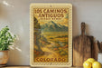A framed vintage-style poster of the scenic route in Los Caminos Antiguo, Colorado, featuring a winding road surrounded by mountains and lush greenery.