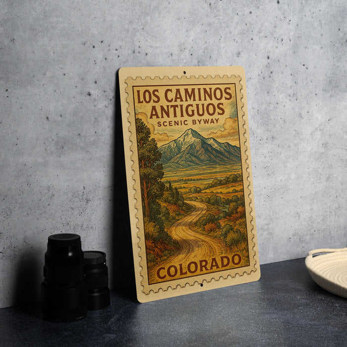 A vintage-style sign for Los Caminos Antiguo, a scenic byway in Colorado, featuring a mountain landscape and a winding road.