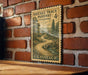 A vintage U.S. postage stamp depicting a winding road through a forest, mounted on a brick wall.