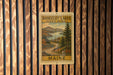 A vintage-style metal sign with a scenic landscape of mountains, trees, and a winding road, mounted on a wooden wall.