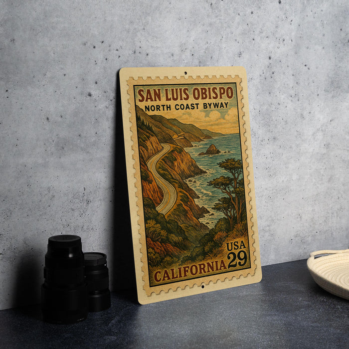A vintage-style metal sign depicting the San Luis Obispo North Coast Byway in California, with a scenic illustration of a winding road and coastal landscape.