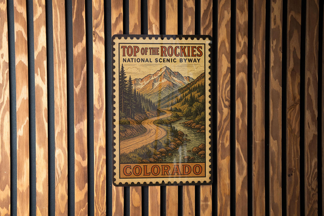 A vintage-style metal sign with a mountain landscape and a winding road, mounted on a wooden wall.