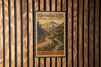 A vintage-style metal sign with a mountain landscape and a winding road, mounted on a wooden wall.