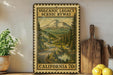 A framed vintage-style stamp depicting a scenic mountain landscape with a winding road, surrounded by pine trees and a lake, is displayed on a shelf next to a wooden cutting board and some lemons.