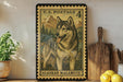 A framed U.S. postage stamp depicting a husky dog, with the text "U.S. POSTAGE 4" and "ALASKAN MALAMUTE" printed on it. The stamp is placed on a shelf next to a wooden cutting board and a potted plant.
