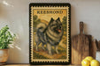 A framed illustration of a dog, specifically a Keeshond, is displayed on a shelf next to a wooden cutting board and some lemons.