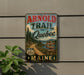 A vintage-style metal sign for the "Arnold Trail to Quebec" in Maine, featuring a scenic road surrounded by pine trees and mountains.